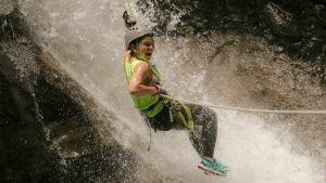 Person in helmet and harness rappelling down a water-drenched rock surface.