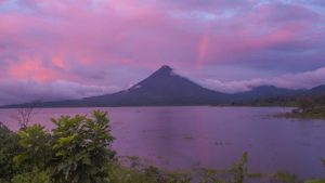 A tranquil lake with a volcano in the background under a pink-hued sunset sky.